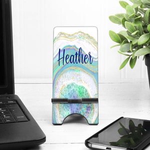 May include: A personalized phone stand with a blue, green, and gold agate design. The name "Heather" is written in blue script across the front. The stand is on a white surface next to a laptop and a smartphone.