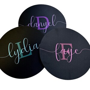 May include: Three black round coasters with personalized names in cursive script. The names are "Danyel" in purple, "Lydia" in teal, and "Faye" in pink. The coasters are arranged in a slight overlap.