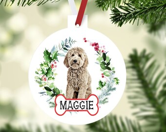 May include: A white Christmas ornament featuring a light brown dog with a floral wreath design. The dog's name, "MAGGIE," is displayed on a bone-shaped graphic. The ornament hangs from a red and white ribbon.