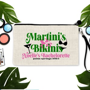 May include: A rectangular, beige cosmetic bag with a black zipper and wrist strap. The bag features the text "Martini's & Bikinis" in green and pink, with a martini glass and bikini graphic. Below the text is "Noelle's Bachelorette palm springs 2024".