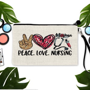 May include: A beige canvas makeup bag with a black zipper and a black strap. The bag has a peace sign, a heart with a leopard print, a stethoscope, a syringe, and the text "Peace. Love. Nursing" in black. The name "Meghan" is written in black above the stethoscope.