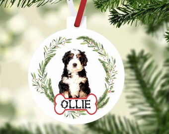 May include: Round white Christmas bauble with a Bernedoodle puppy illustration. The puppy is framed by a green wreath with red berries. The name "OLLIE" is on a red bone-shaped tag. A red and white ribbon is attached for hanging on the tree.