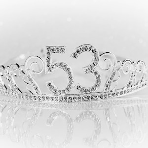 May include: A silver tiara with the number "53" encrusted with rhinestones. The tiara has an ornate design with swirls and is reflected on a white surface. The image is in black and white.