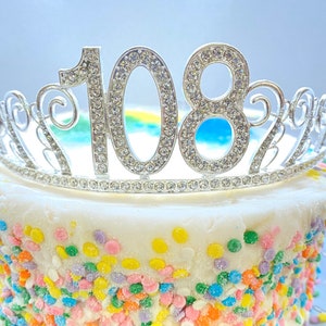 May include: A white cake with colourful sprinkles topped with a silver tiara featuring the number 108 in rhinestones.
