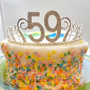 May include: A white cake decorated with colorful sprinkles and topped with a silver tiara with the number 59 on it.