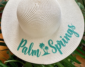 May include: A cream-colored sun hat with a wide brim. The hat features the text "Palm Springs" in teal glitter script, along with a palm tree graphic. The hat is made of woven material and is suitable for sun protection.