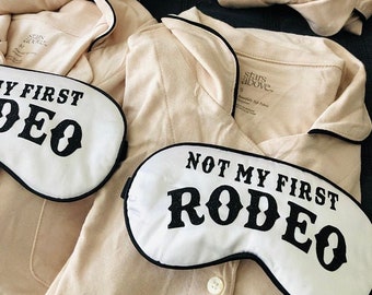 May include: Two white sleep masks with black trim and the text "Not My First Rodeo" and "My First Rodeo". The masks are on top of beige pajamas with black trim.