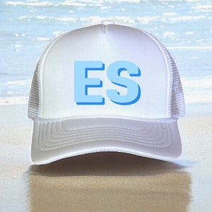 May include: White trucker hat with a blue "ES" logo on the front. The hat is sitting on a sandy beach with the sea in the background.