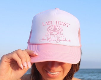 May include: A pink and white trucker hat with the text "Last Toast 2024 On The Coast Anna Marie's Bachelorette" in pink lettering. The hat features a white shell design.