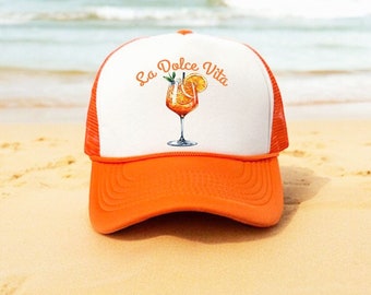May include: Orange and white trucker hat with a white mesh back. The hat has an orange brim and a white front with the text "La Dolce Vita" and a drawing of an orange drink with a slice of orange.