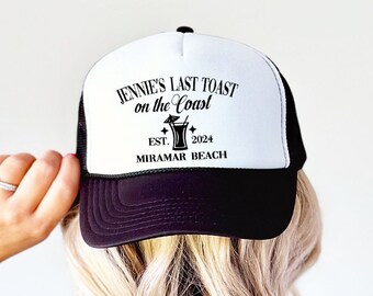 May include: A black and white trucker hat featuring the text "Jennie's Last Toast on the Coast" and a cocktail illustration. The hat also includes "Est. 2024 Miramar Beach". The hat has a black brim and mesh back.