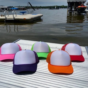 May include: Five trucker hats with white fronts and mesh backs in various colors: pink, purple, green, orange, and red. The hats are displayed on a dock with a lake and boats in the background.