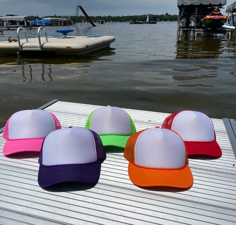 May include: Five trucker hats with white fronts and mesh backs in various colors: pink, purple, green, orange, and red. The hats are displayed on a dock with a lake and boats in the background.