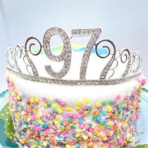 Peut inclure: Une tiare argentée avec des strass qui épellent le nombre 1977 est posée sur un gâteau blanc décoré de vermicelles colorés.
