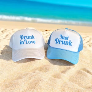 May include: Two white and blue trucker hats with mesh backs. One hat says "Drunk in Love" in blue lettering and the other says "Just Drunk" in blue lettering. The hats are on a sandy beach.