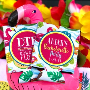 May include: Two white foam beverage insulators featuring a tropical pattern of flamingos, palm leaves, and fruit. One displays the text DTF DOWN TO FLAMINGO, while the other reads AMBER'S Bachelorette Party 8-24-19 inside a circular magenta frame.