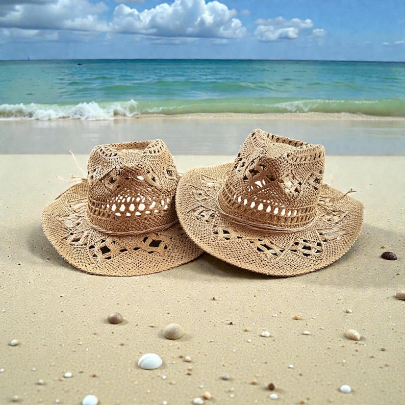 K&ouml;nnte beinhalten: Zwei gewebte Strohh&uuml;te mit dekorativen Ausschnitten liegen an einem Sandstrand. Die H&uuml;te haben eine klassische Cowboyhutform mit breiter Krempe und leicht eingedellter Krone. Der Hintergrund zeigt einen t&uuml;rkisfarbenen Ozean und einen blauen Himmel mit Wolken.