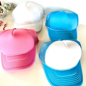 May include: Four trucker hats in various colors: white, light blue, pink, and a blue and white combination. The hats have a foam front and a mesh back, with a curved brim. The hats are displayed on a white surface.