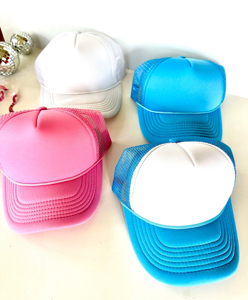May include: Four trucker hats in various colors: white, light blue, pink, and a blue and white combination. The hats have a foam front and a mesh back, with a curved brim. The hats are displayed on a white surface.