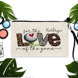 May include: A beige zippered makeup bag with a black zipper and a black strap. The bag has a golf ball with a red heart on it and the words "for the love of the game" in black script. The name "Kathryn" is written in black script above the golf ball.