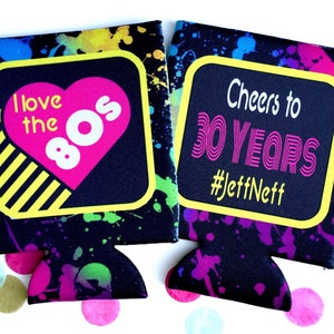 May include: Two black neoprene can coolers with a colorful paint splatter design. One cooler says "I love the 80s" with a pink heart and the other says "Cheers to 30 Years #JeffNeff".