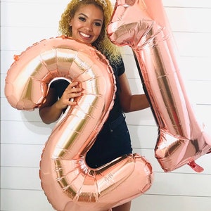 May include: A person holding two large rose gold foil balloons shaped like the numbers 2 and 1. The balloons are in front of a white wood plank wall.