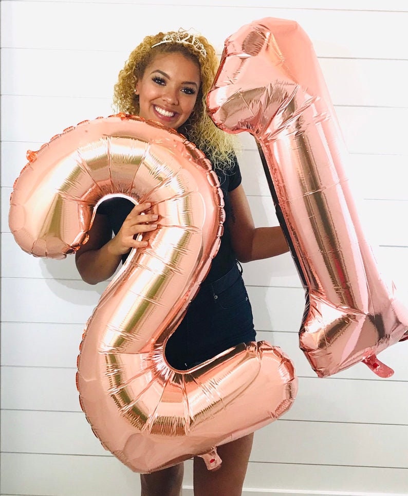 May include: A person holding two large rose gold foil balloons shaped like the numbers 2 and 1. The balloons are in front of a white wood plank wall.