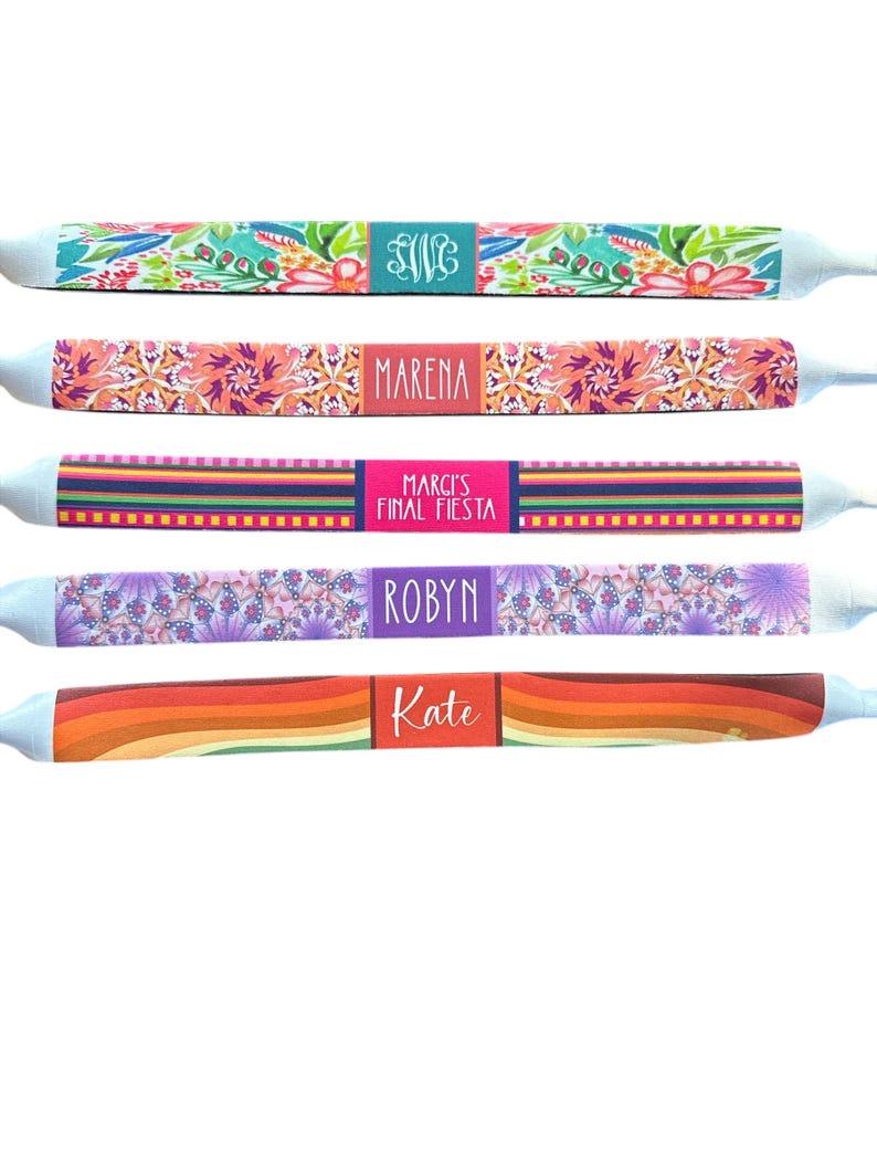 May include: A collection of five personalized eyeglass straps. Each strap features a unique design, including floral patterns, stripes, and a rainbow gradient. Names or initials are displayed on each strap, such as "SWG", "Marena", "Robyn", and "Kate".