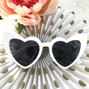 May include: White heart-shaped sunglasses with black lenses. The sunglasses are on a white and gold background.