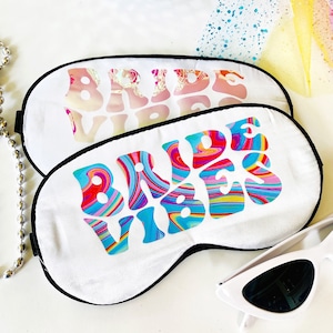 May include: Two white sleep masks with black trim. One mask has iridescent text that reads "BRIDE VIBES". The other mask has colorful, swirling text that reads "BRIDE VIBES". A pair of white sunglasses is next to the masks.