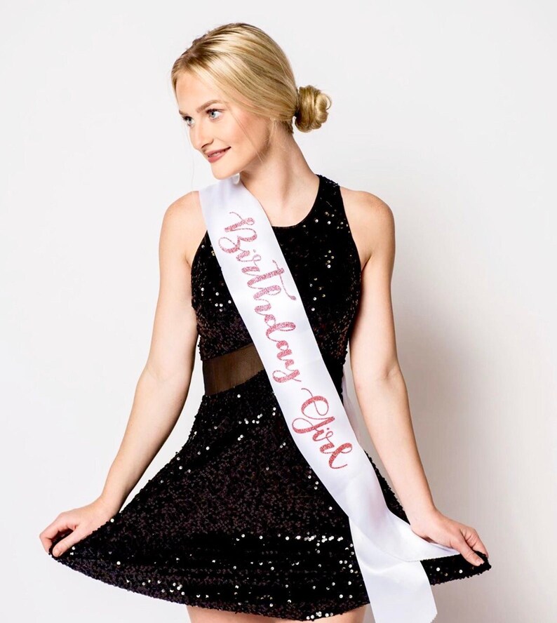 May include: A white satin sash with the words "Birthday Girl" written in pink glitter. The sash is worn by a person wearing a black sequined dress.