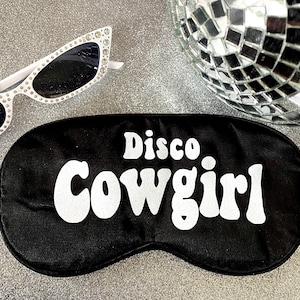 May include: Black sleep mask with the words "Disco Cowgirl" in white, retro-style lettering. White sunglasses with rhinestone accents are in the background, along with a disco ball.