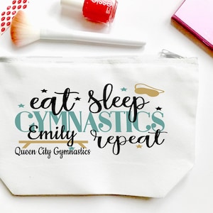 May include: White canvas cosmetic bag with a zipper closure. The bag features the text "eat sleep gymnastics Emily repeat" in black and teal lettering, with a gold-colored gymnastics bar and eye mask graphic. Additional makeup items are visible.