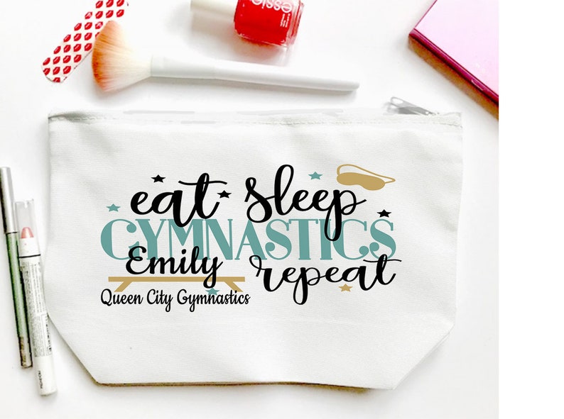May include: White canvas cosmetic bag with a zipper closure. The bag features the text "eat sleep gymnastics Emily repeat" in black and teal lettering, with a gold-colored gymnastics bar and eye mask graphic. Additional makeup items are visible.