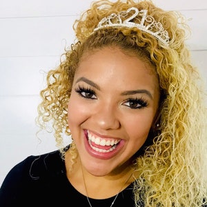 May include: A smiling person with curly blonde hair wearing a silver tiara with the number "21" on it. The person is wearing a black top and a silver necklace. The background is a white wall.