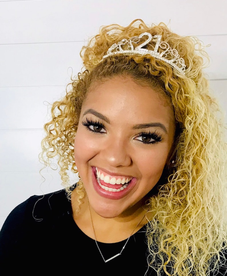 May include: A smiling person with curly blonde hair wearing a silver tiara with the number "21" on it. The person is wearing a black top and a silver necklace. The background is a white wall.