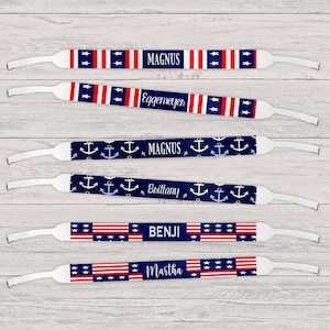 May include: Several pairs of eyeglass straps with patriotic designs. Some straps feature red and white stripes with blue stars, while others have a navy blue background with white anchors. Names like "Magnus," "Benji," and "Martha" are printed on the straps.