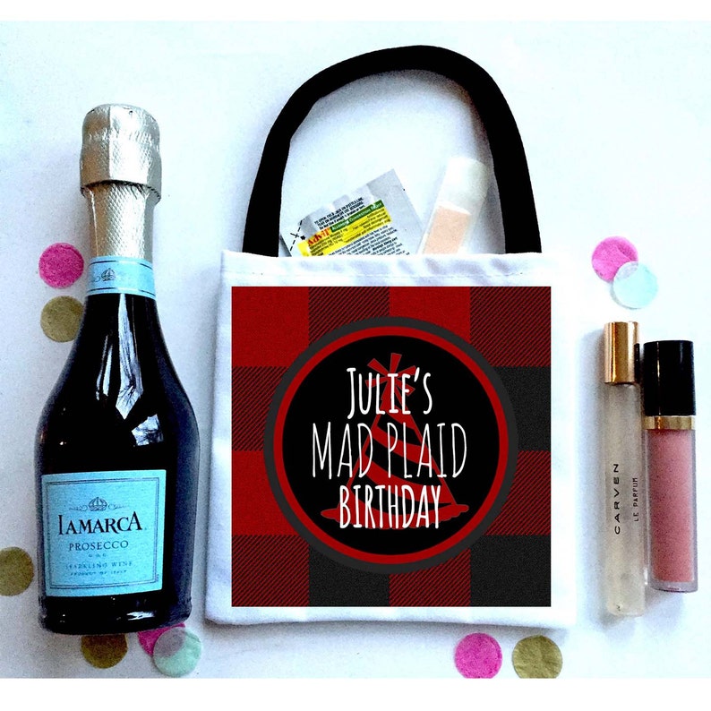 May include: A white canvas tote bag with black handles. The bag has a red and black plaid pattern and a circle with the text "Julie's Mad Plaid Birthday".