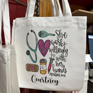 May include: White canvas tote bag with a colourful design featuring a stethoscope, heart, bandage, pill bottle, and syringe. The text reads "She works willingly with her hands Proverbs 31:13" and "Courtney".