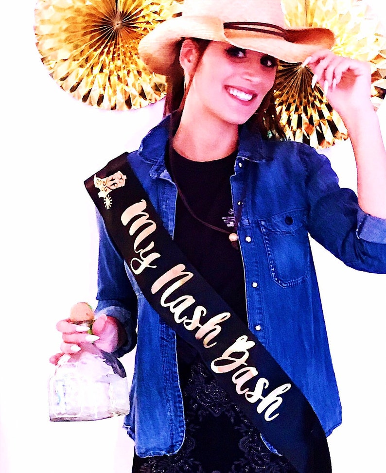 Puede incluir: Una mujer con una camisa vaquera y un sombrero de vaquero sostiene una botella de tequila. Lleva una banda negra con letras doradas que dicen "My Mash Bash".