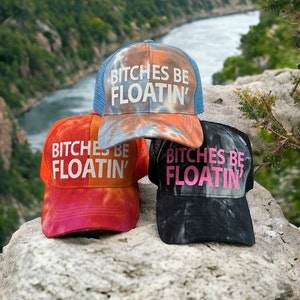 May include: Three tie-dye trucker hats with the phrase "Bitches Be Floatin'" in white, pink, and white lettering. The hats are on a rock with a river in the background.