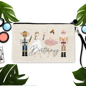 May include: A beige canvas makeup bag with a black zipper and a black strap. The bag features a colorful illustration of a ballerina, a nutcracker, and a mouse king. The name "Brittany" is written in cursive on the bag.