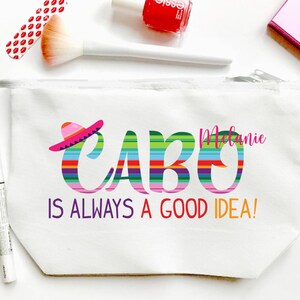 May include: White canvas cosmetic bag with the text "CABO IS ALWAYS A GOOD IDEA!" in colourful letters. The word "CABO" has a striped design and a sombrero. The name "Melanie" is written in pink. Makeup brushes and nail polish are visible.