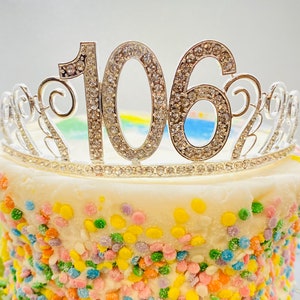 May include: A white cake decorated with colorful sprinkles and topped with a silver tiara featuring the number "106" in rhinestones.