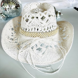 May include: White crochet cowboy hat with a pearl and rhinestone trim. The hat has a wide brim and a crown with a lacy design.