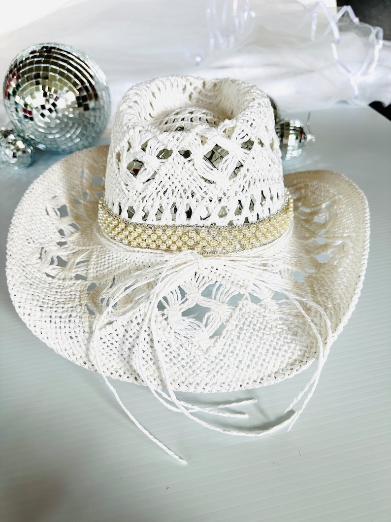 May include: White crochet cowboy hat with a pearl and rhinestone trim. The hat has a wide brim and a crown with a lacy design.