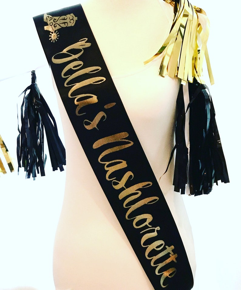 Puede incluir: Sash de sat&eacute;n negro con letras doradas que dicen "Bella's Bachelorette" y una bota dorada con purpurina y una espuela.