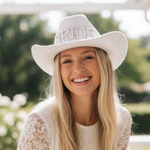 K&ouml;nnte beinhalten: Ein wei&szlig;er Cowboyhut mit dem Wort "BRIDE" in silbernen Strasssteinen geschrieben. Der Hut hat eine gebogene Krempe und eine abgerundete Krone. Ein Brautaccessoire.