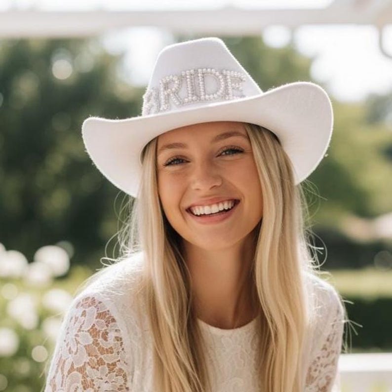 K&ouml;nnte beinhalten: Ein wei&szlig;er Cowboyhut mit dem Wort "BRIDE" in silbernen Strasssteinen geschrieben. Der Hut hat eine gebogene Krempe und eine abgerundete Krone. Ein Brautaccessoire.