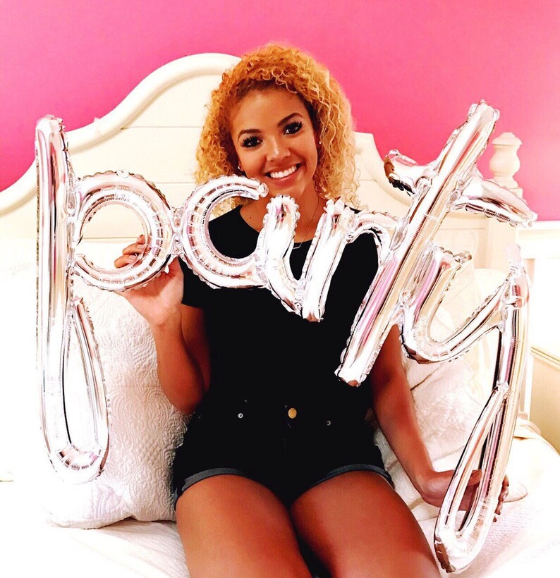 May include: A person holding silver foil balloons that spell out "party" in front of a pink wall.